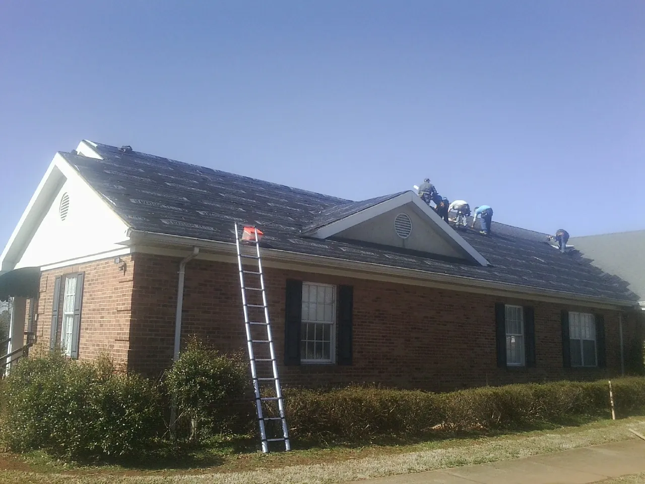 Crew tearing shingles off a brick ranch roof with a ladder set — Emergency Roof Repair work by our crew in Quenemo