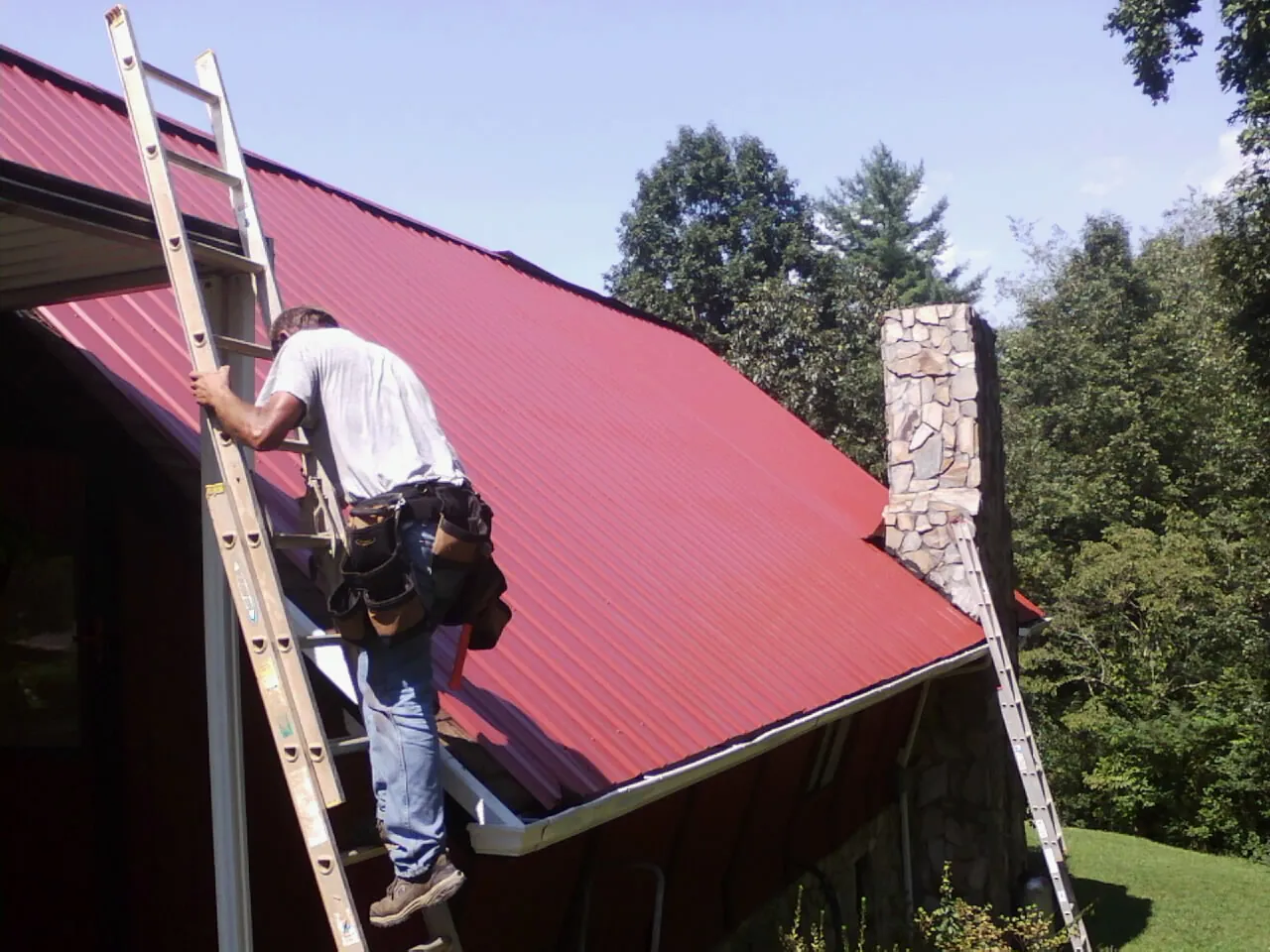 Roofer on a ladder beside a red metal roof and stone chimney — Soffit & Fascia Repair by Roofing Brothers in Quenemo